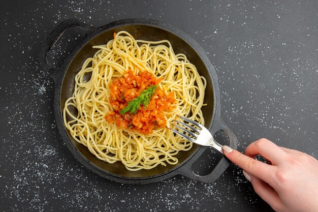 Вид сверху спагетти с соусом на сковороде, вилка в женской руке на черном столе, свободное пространство