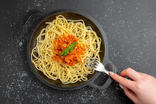 Вид сверху спагетти с соусом на сковороде, вилка в женской руке на черном столе, свободное пространство