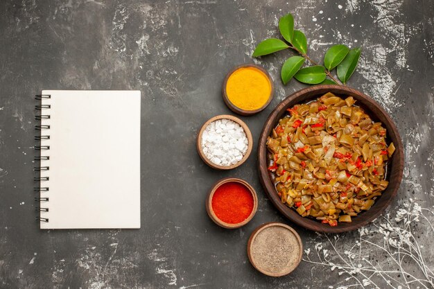 Top view spices plate of green beans next to the bowls of colorful spices leaves next to the white notebook and tree branches on the dark table