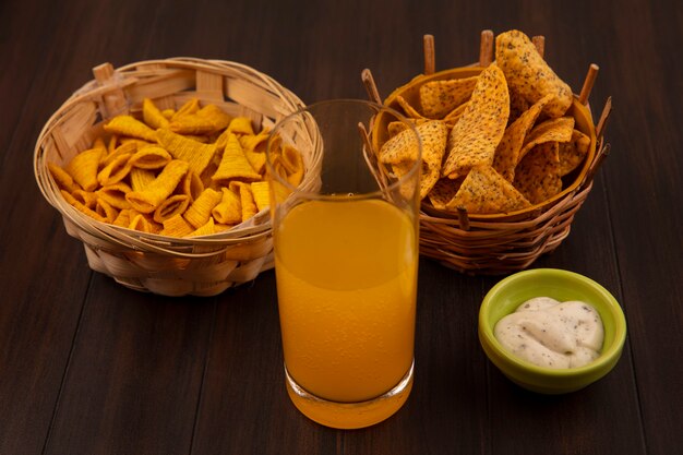 木製のテーブルの上の緑のボウルにソースとオレンジジュースのガラスとバケツの上のトウモロコシのスナックとバケツのスパイシーなチップの上面図