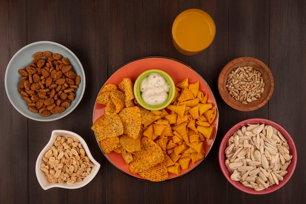 Top view of spicy crispy chips on an orange dish with sauce on a bowl with shelled sunflower seeds on a wooden bowl with pine nuts on a bowl with a glass of orange juice on a wooden table