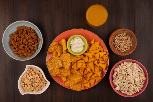 Top view of spicy crispy chips on an orange dish with sauce on a bowl with shelled sunflower seeds on a wooden bowl with pine nuts on a bowl with a glass of orange juice on a wooden table