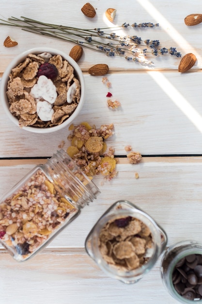 Top view of spilled granola jar near cornflakes; dry fruits and chocolate chips on wooden surface