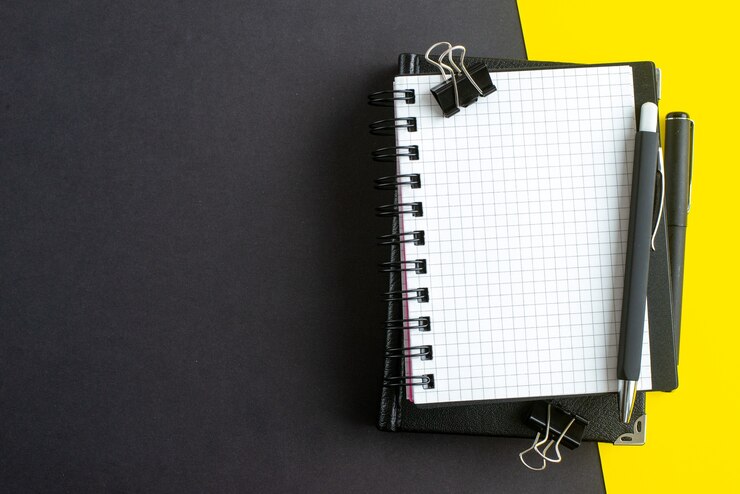 Free Photo top view of spiral notebook on book and pens on black yellow background with free space