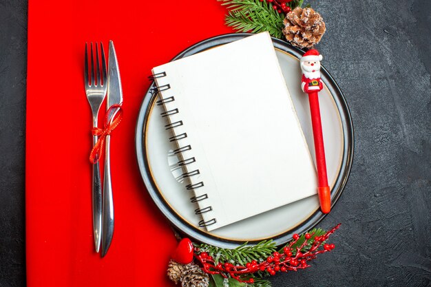 Top view of a spiral notebook and a pen on dinner plate with decoration accessories fir branches and cutlery set on a red napkin