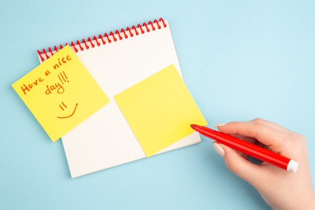 Top view spiral notebook red marker in woman hand have a nice day written on yellow note paper on blue