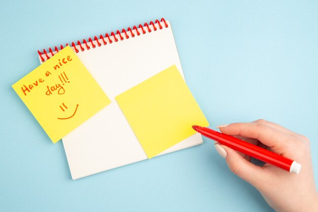 Вид сверху спиральный блокнот, красный маркер в руке женщины, хорошего дня, написанного на желтой бумаге для заметок на синем