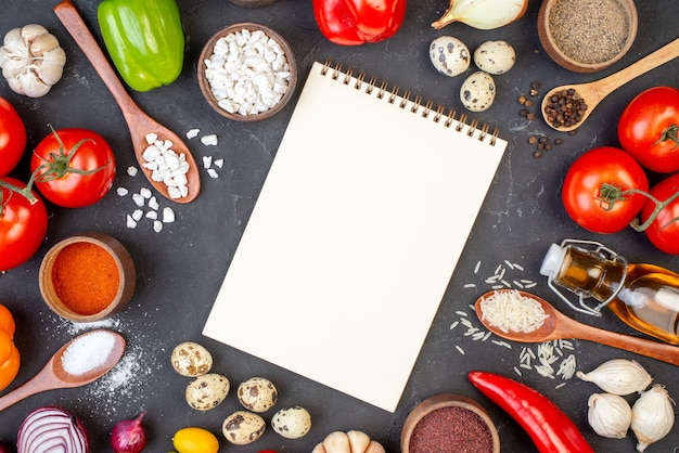 Top view spiral notebook rice sea salt in wooden spoons tomatoes quail eggs cumcuat garlic oil bottle on black table