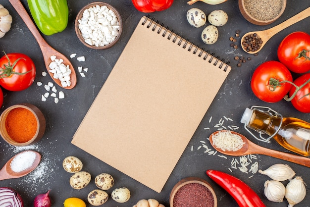 Top view spiral notepad rice sea salt in wooden spoons tomatoes quail eggs oil bottle spices in bowls onions on black table