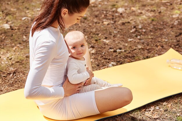 Top view of sporty woman with infant kid sitting on karemat in lotus pose, keeping legs crossed