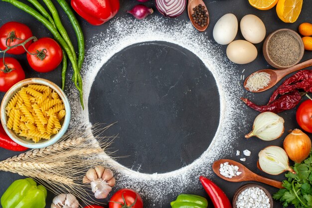 Top view sprinkled flour circle tomatoes garlic eggs spiral pasta in bowl bell peppers wooden spoons on table free space