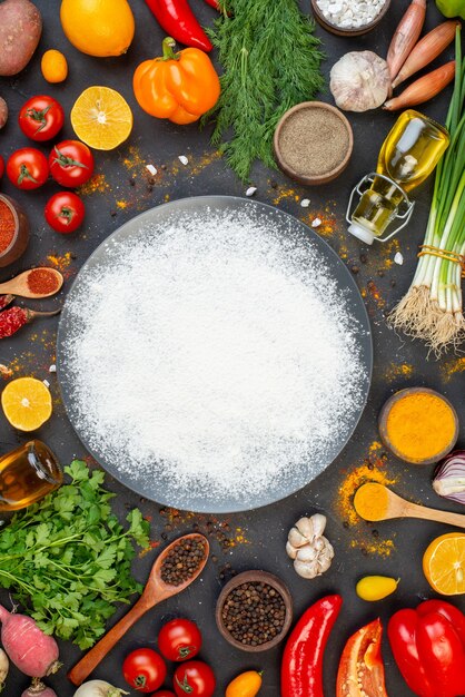 Вид сверху рассыпанной мукой на круглой тарелке, порошка черного перца и куркумы в небольших мисках, кориандра, помидоров, чеснока, перца, лимона на столе