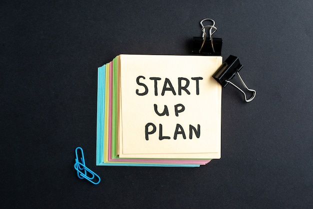 Top view of START UP PLAN writing on small note sheets simple office tools on black background with free space