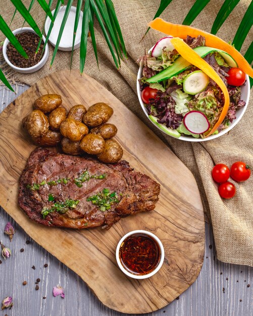 Top view steak with potatoes and sauce on the board with salad and vegetables