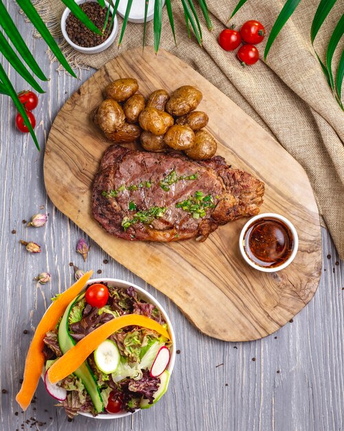 Top view steak with potatoes and sauce on the board with salad and vegetables