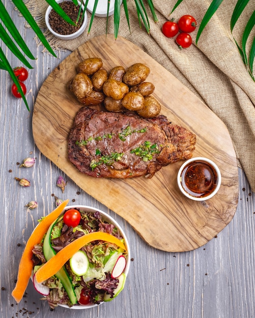 Top view steak with potatoes and sauce on the board with salad and vegetables