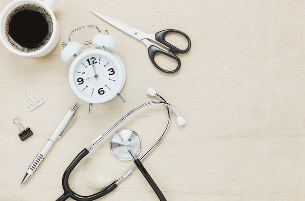 Top view stethoscope with clock coffee scissors with pen on wooden background.