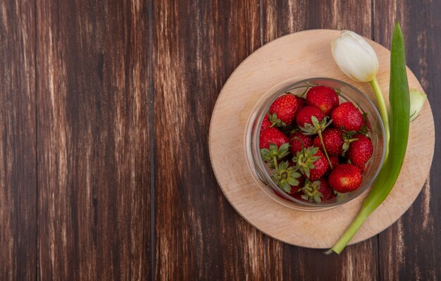 Top view of strawberries in bowl and flower on cutting board on wooden background with copy space