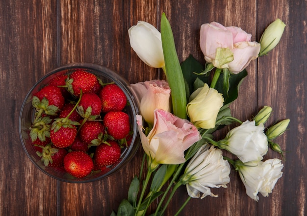 Top view of strawberries in bowl and flowers on wooden background