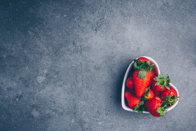Top view strawberries in heart shaped bowl on gray textured background. horizontal free space for your text