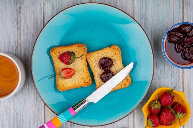 Top view of strawberry on bread on a blue plate with knife with strawberry jam on a bowl with fresh strawberries on a grey wooden background