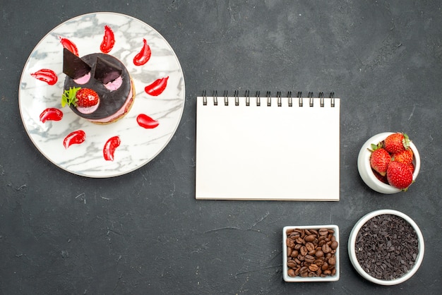 Top view strawberry cheesecake on oval plate bowls with strawberries chocolate coffee seeds a notepad on dark surface