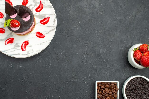 왼쪽 상단의 흰색 타원형 접시에 있는 상단 뷰 딸기 치즈 케이크와 어두운 표면의 오른쪽 하단에 딸기 초콜릿 커피 씨앗이 있는 그릇
