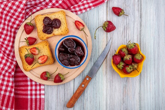 Top view of strawberry jam on a bowl with fresh strawberries on a wooden kitchen board with knife with fresh strawberries on a yellow bowl on a grey wooden background