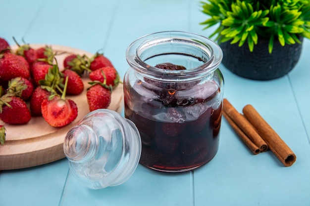 Top view of strawberry jam on a glass jar with cinnamon sticks with fresh strawberries on a wooden kitchen board on a blue background