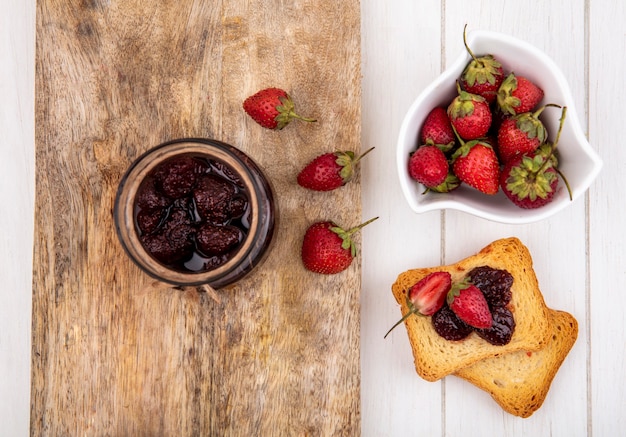 Top view of strawberry jam on a glass jar on a wooden kitchen board with fresh strawberries on a white bowl with toasted slices of bread on a white wooden background