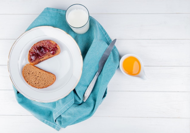 Top view of strawberry jam smeared on sliced rye bread in plate with knife milk on blue cloth and melted butter on wooden table with copy space