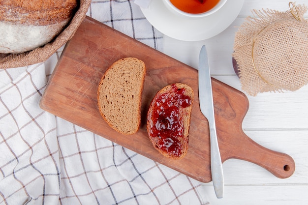 Top view of strawberry jam smeared on sliced rye bread with knife on cutting board and cob on plaid cloth with tea and jam on wooden table
