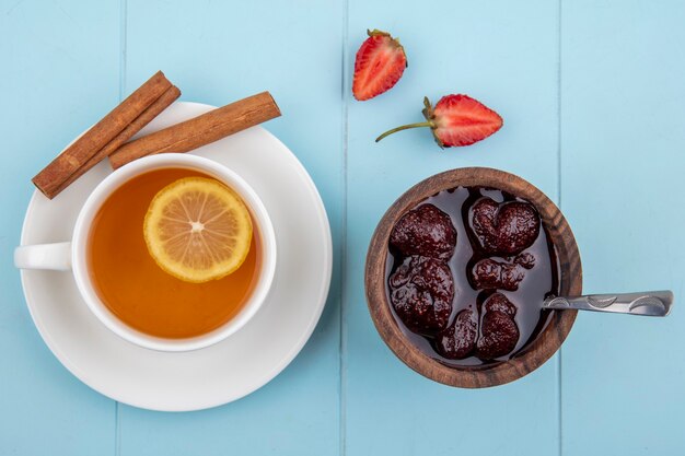 Top view of a strawberry jam on a wooden bowl with teaspoon with a cup of tea with cinnamon on a blue background