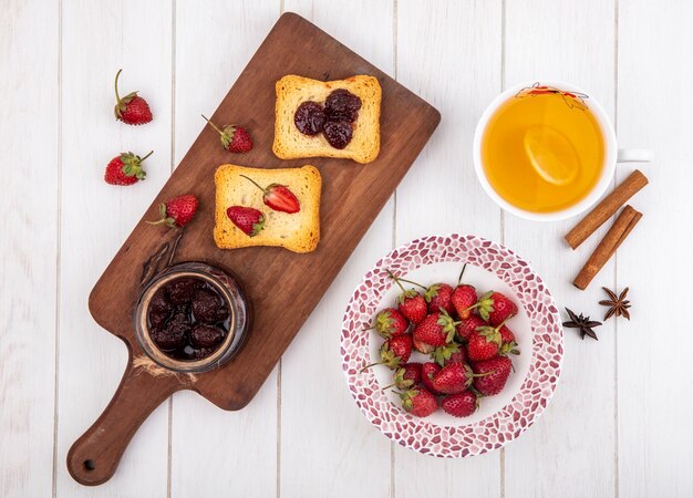 Top view of strawberry jam on a wooden kitchen board with toasted bread with fresh strawberries with cinnamon sticks on a white wooden background