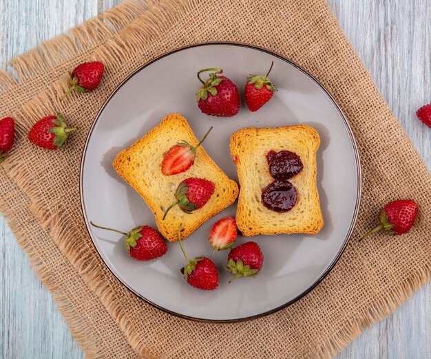 Top view of strawberry on toasted bread on a plate with fresh strawberries isolated on a sack cloth on a grey wooden background