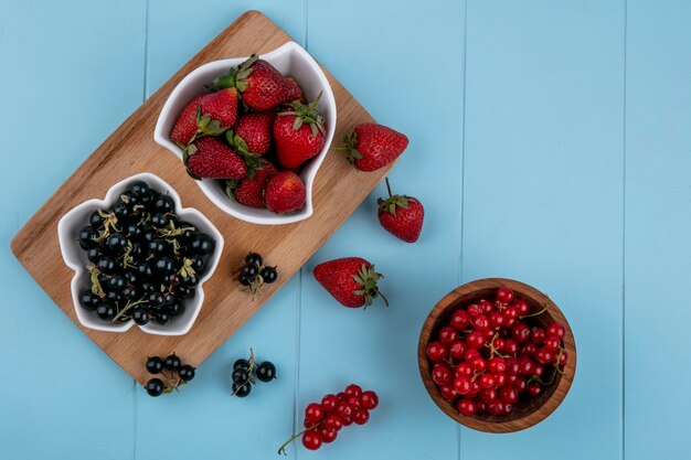 Top view strawberry with black currants on a board with red currants in a bowl on a blue background