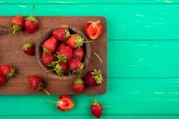 Top view of strawberry on a wooden bowl on a wooden kitchen board on a green background with copy space