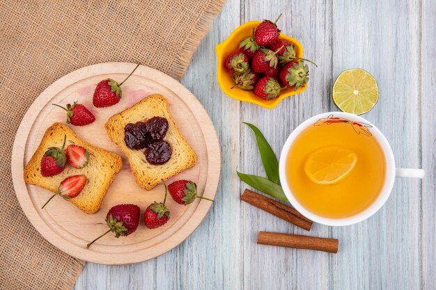 Top view of strawberry on a wooden kitchen board on sack cloth with cinnamon sticks with a cup of tea on a grey wooden background