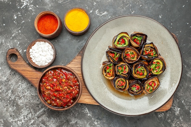 Top view stuffed aubergine rolls in oval plate adjika in bowl on wooden serving board with handle different spices in small bowls on grey background