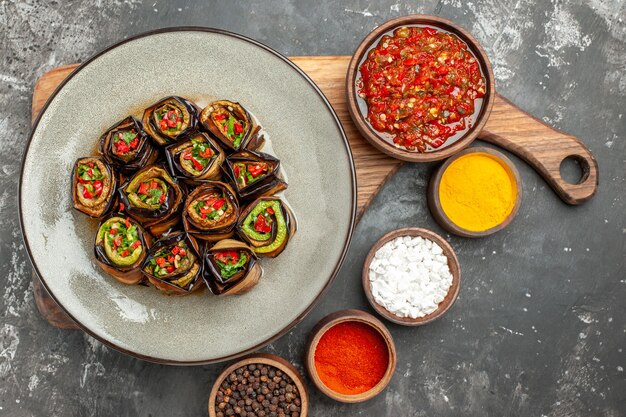 Top view stuffed aubergine rolls in white oval plate adjika in bowl on wooden serving board with handle different spices in small bowls on grey background