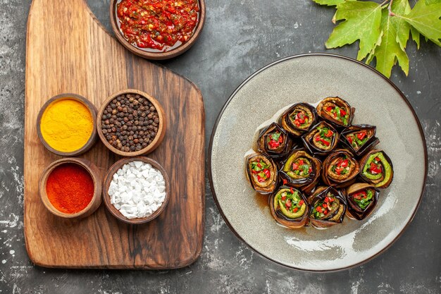 Top view stuffed aubergine rolls in white oval plate different spices in bowls on wooden serving board with handle adjika on grey background