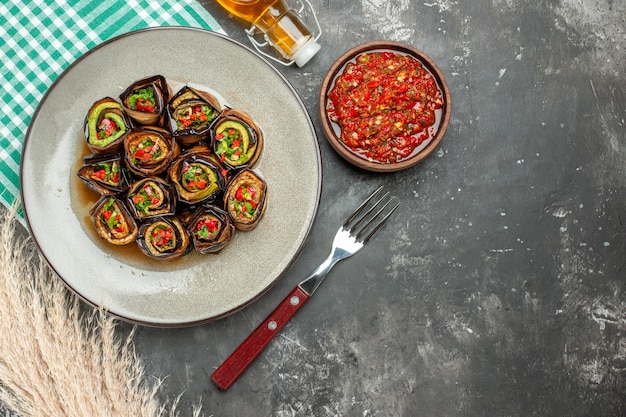 Top view stuffed aubergine rolls in white oval plate a fork bottle of oil adjika on grey surface