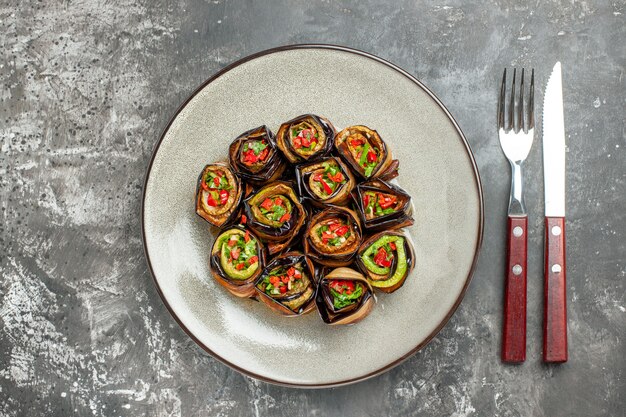 Top view stuffed aubergine rolls on white oval plate fork and knife on grey surface