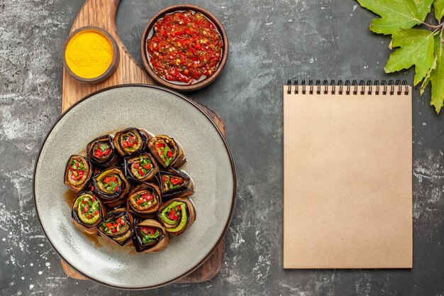 Top view stuffed aubergine rolls in white oval plate turmeric in bowl on wooden serving board with handle adjika a notebook on grey background