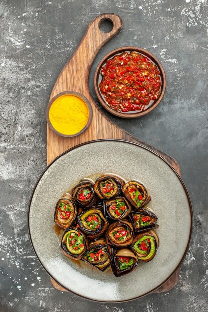 Top view stuffed aubergine rolls in white oval plate turmeric in bowl on wooden serving tray with handle adjika on grey surface