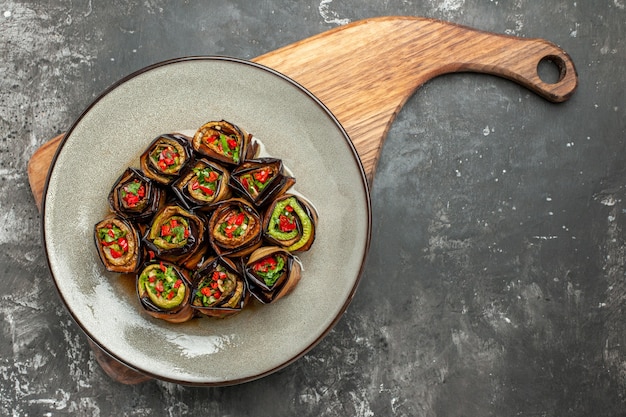 Top view stuffed aubergine rolls in white oval plate on wooden serving board with handle on grey background