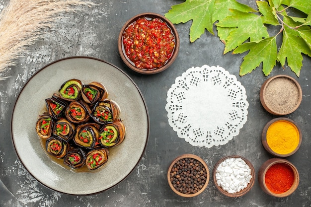 Top view stuffed eggplant rolls different spices adjika in small bowls white oval lace doily on grey background