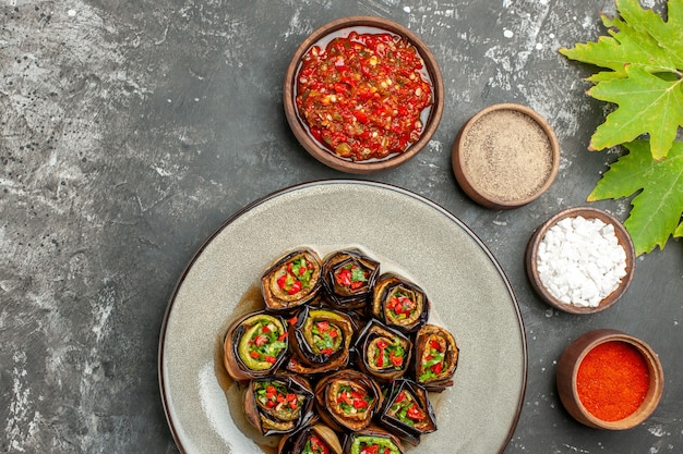 Top view stuffed eggplant rolls in white oval plate spices in small bowls salt pepper red pepper adjika on grey surface