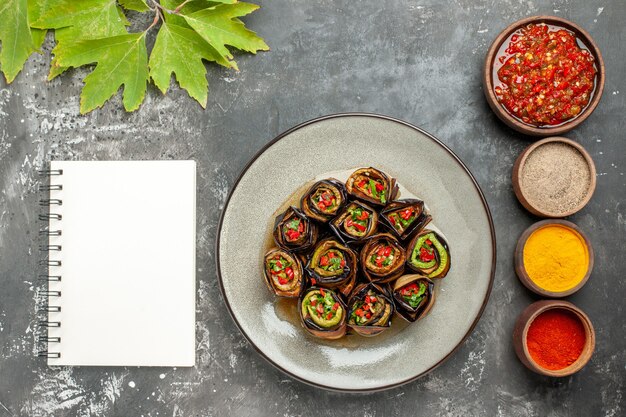 Top view stuffed eggplant rolls in white plate different spices adjika in small bowls a notebook on grey surface