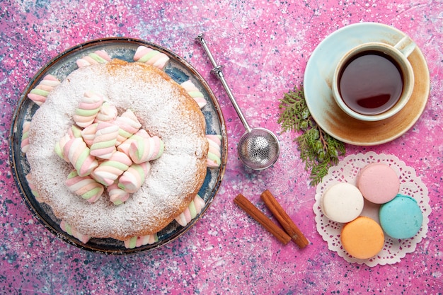 Вид сверху сахарного торта с чашкой чая и макаронами на розовой поверхности